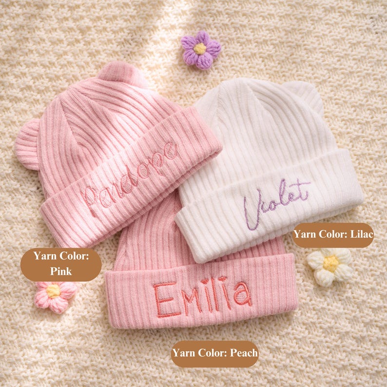 May include: Three knitted baby beanies in pink and white, with embroidered names and small ear details. The pink beanies have the names "Penelope" and "Emilia", while the white beanie is embroidered with "Violet". Decorative yarn flowers are also present.