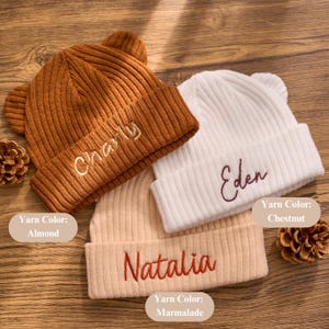 May include: Three ribbed knit beanies with bear ears, each embroidered with a name. Colors include almond, chestnut, and marmalade. The names embroidered are Charly, Eden, and Natalia. The beanies are a cozy accessory.