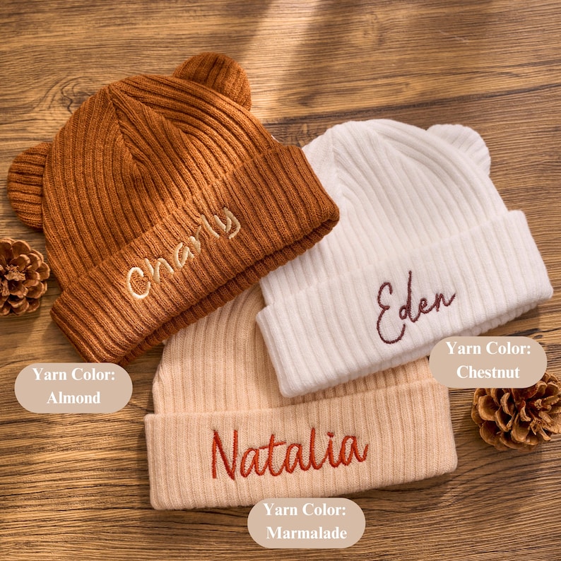 May include: Three ribbed knit beanies with bear ears, each embroidered with a name. Colors include almond, chestnut, and marmalade. The names embroidered are Charly, Eden, and Natalia. The beanies are a cozy accessory.