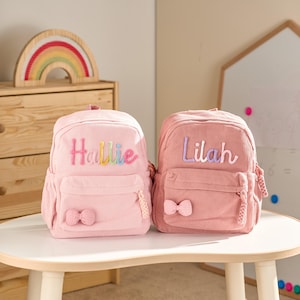 May include: Two pink corduroy backpacks with embroidered names, Hallie and Lilah, in colorful and lavender thread. Each backpack has a front pocket with a bow and a braided zipper pull. A wooden rainbow decorates the background.