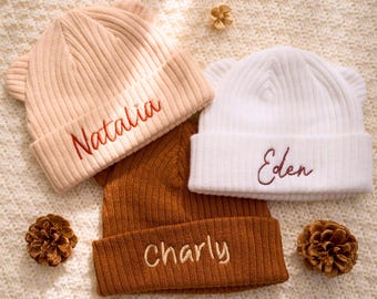 May include: Three personalized knit beanies in beige, white, and brown, each with embroidered names: "Natalia," "Eden," and "Charly." The hats feature small ear details and are arranged on a textured, off-white surface with pinecones.