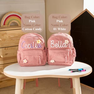 May include: Two pink corduroy backpacks featuring embroidered names and small flower appliqu&eacute;s. The left bag displays the name Cecilia in light purple, and the right bag displays the name Bella in white. Text labels indicate bag and name colours.