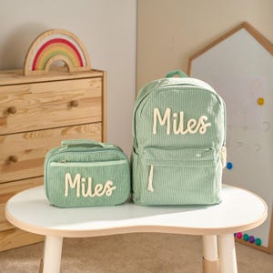May include: A light green corduroy backpack and matching lunch bag, both with the name "Miles" in white script. The backpack has a front pocket and adjustable straps. The lunch bag has a handle and a zipper closure.