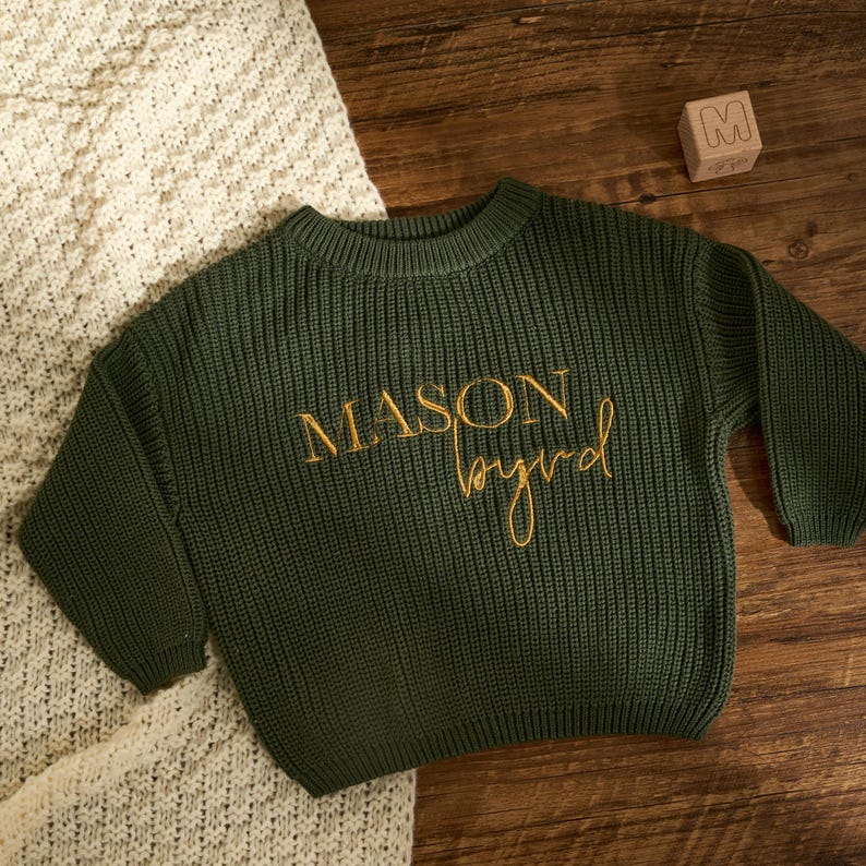 May include: An olive green knitted sweater with the name "MASON byrd" embroidered in gold thread. The sweater is on a wooden surface next to a cream knitted blanket and a wooden block with the letter "M".