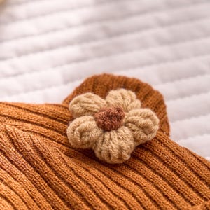 May include: Close-up of a knitted, rust-colored garment with a ribbed texture. A beige and brown crocheted flower is attached. The flower has five petals and a darker brown center, adding a decorative touch to the piece.