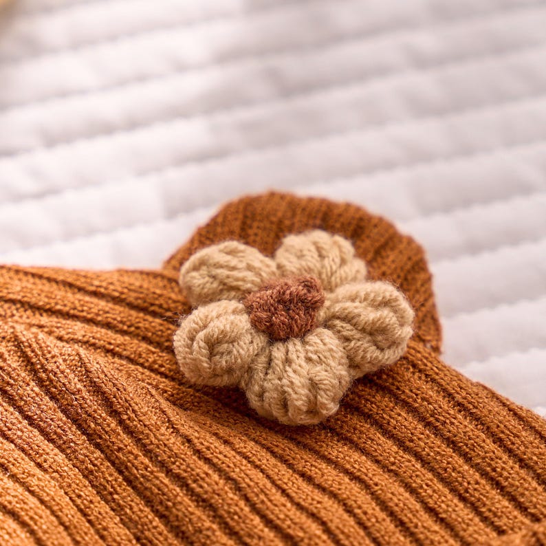 May include: Close-up of a knitted, rust-colored garment with a ribbed texture. A beige and brown crocheted flower is attached. The flower has five petals and a darker brown center, adding a decorative touch to the piece.