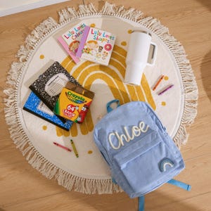 May include: A light blue corduroy backpack with the name "Chloe" embroidered on it, along with a rainbow patch. The backpack is surrounded by school supplies, including notebooks, crayons, and children's books, all on a round rug.