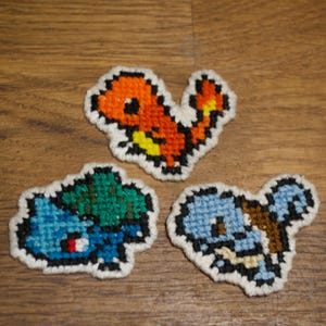 May include: Three embroidered patches featuring Pokemon characters. The patches are made with colourful yarn and depict Charmander, Squirtle, and Bulbasaur.