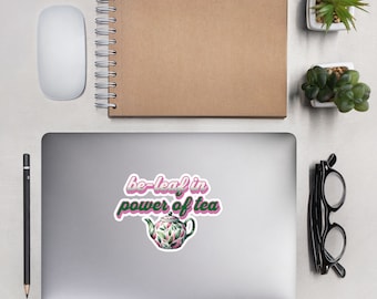 Könnte beinhalten: Ein Aufkleber mit dem Text "be-leaf in power of tea" in Rosa und Grün. Der Aufkleber zeigt eine Teekannen-Illustration mit grünen Blättern und rosa Akzenten. Der Aufkleber befindet sich auf einem Laptop, mit einer Maus, einem Notizbuch, einem Stift, einer Brille und kleinen Pflanzen in der Nähe.