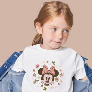 May include: A young girl wearing a white t-shirt featuring a Minnie Mouse graphic with a pink bow and autumn-themed elements. The design includes leaves, acorns, and flowers in warm colors.