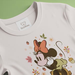 May include: Cream-colored t-shirt featuring a Minnie Mouse graphic with an autumn theme. The design includes a brown bow, leaves, acorns, and flowers. The shirt has a tag that reads "Enarmonia".