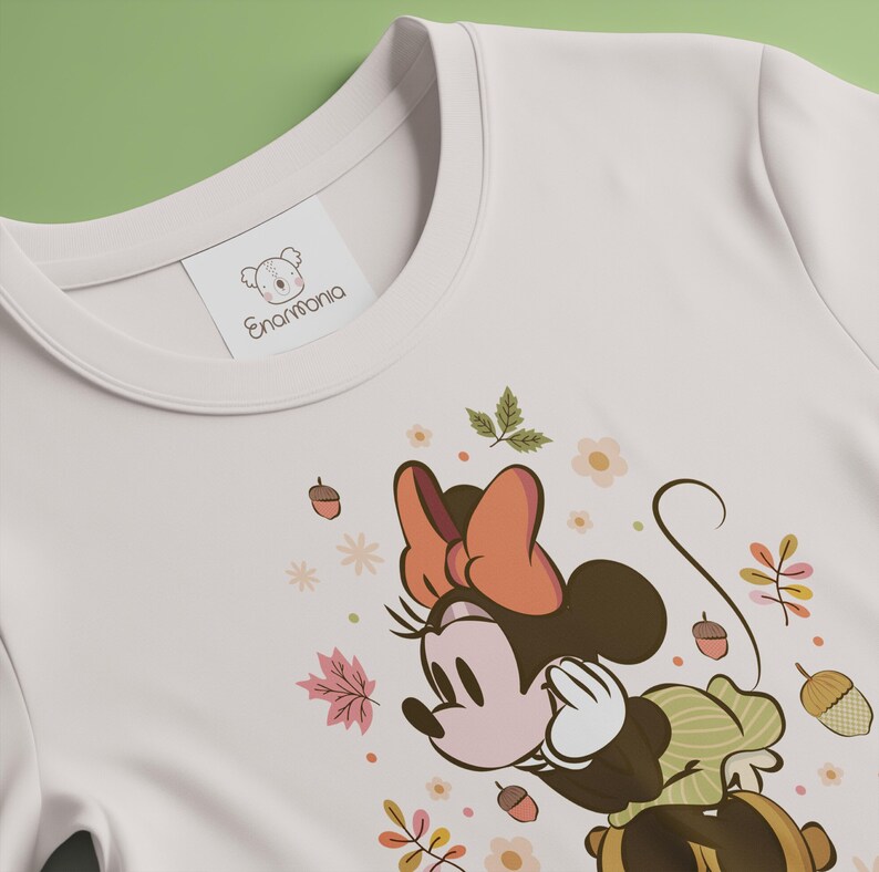 May include: Cream-colored t-shirt featuring a Minnie Mouse graphic with an autumn theme. The design includes a brown bow, leaves, acorns, and flowers. The shirt has a tag that reads "Enarmonia".