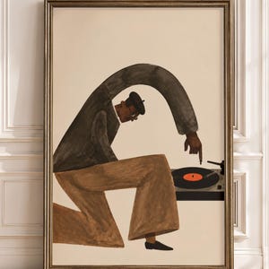 Op de afbeelding: Een ingelijste muurkunstprint met een gestileerde illustratie van een persoon in een donkere trui, bruine broek en een baret. De figuur knielt en gebruikt een verlengde arm om een naald op een zwarte vinylplaat op een platenspeler te plaatsen.