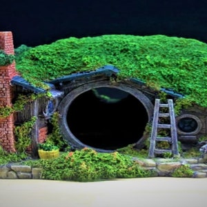 Può includere: Una casa di hobbit ricoperta di muschio verde con un camino in mattoni, una porta rotonda e una scala di legno. La casa è realizzata in resina marrone e grigia ed è un pezzo decorativo per un acquario o un terrario.