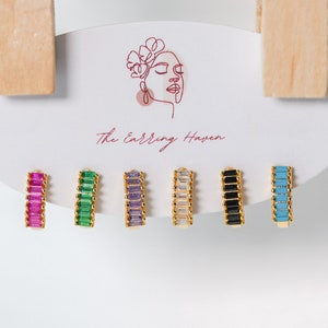 May include: Six pairs of gold hoop earrings with different colored crystal stones. The earrings are displayed on a white card with the text "The Earring Haven".