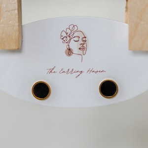 May include: A pair of black stud earrings with gold settings. The earrings are displayed on a white card with the text "The Earring Haven".