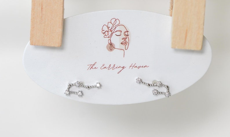 May include: A pair of silver constellation earrings with small, clear stones. The earrings are displayed on a white card with the text "The Earring Haven".