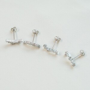 May include: Four silver stud earrings with a linear design, each set with small, clear gemstones. The earrings are displayed on a white surface, highlighting their delicate and sparkling appearance. The studs are designed for pierced ears.