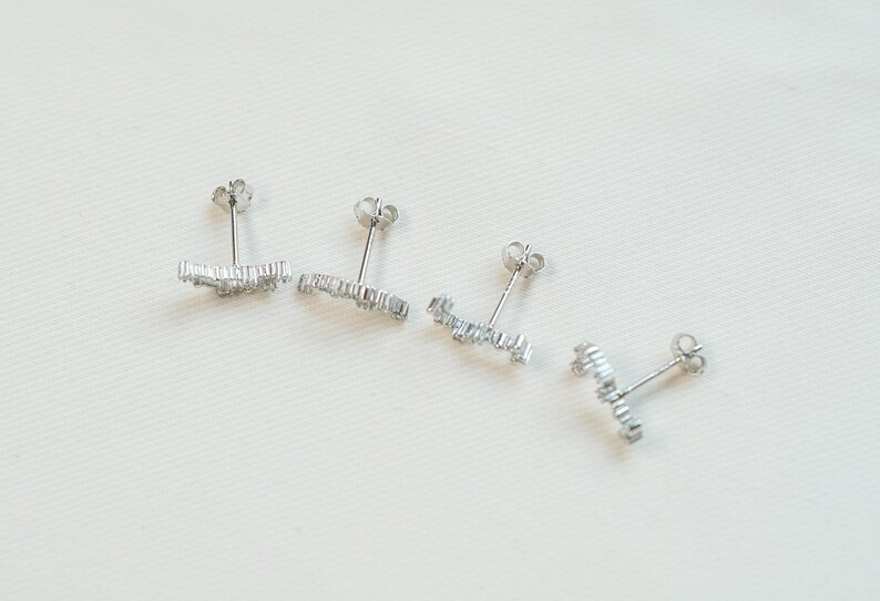 May include: Four silver stud earrings with a linear design, each set with small, clear gemstones. The earrings are displayed on a white surface, highlighting their delicate and sparkling appearance. The studs are designed for pierced ears.
