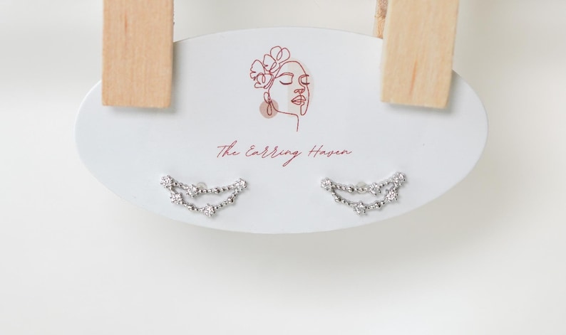 May include: Silver-coloured earrings with a curved design, featuring small, sparkling stones. The earrings are presented on a white oval card with the text "The Earring Haven" and a line drawing of a woman's face.