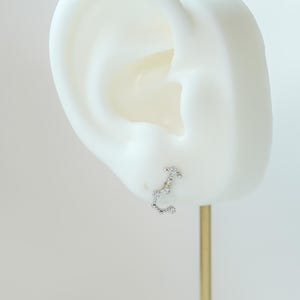 May include: A silver stud earring shaped like the Scorpio constellation, adorned with small, clear gemstones. The earring is presented on a white ear-shaped display against a pale backdrop. The earring is attached to a gold-coloured post.
