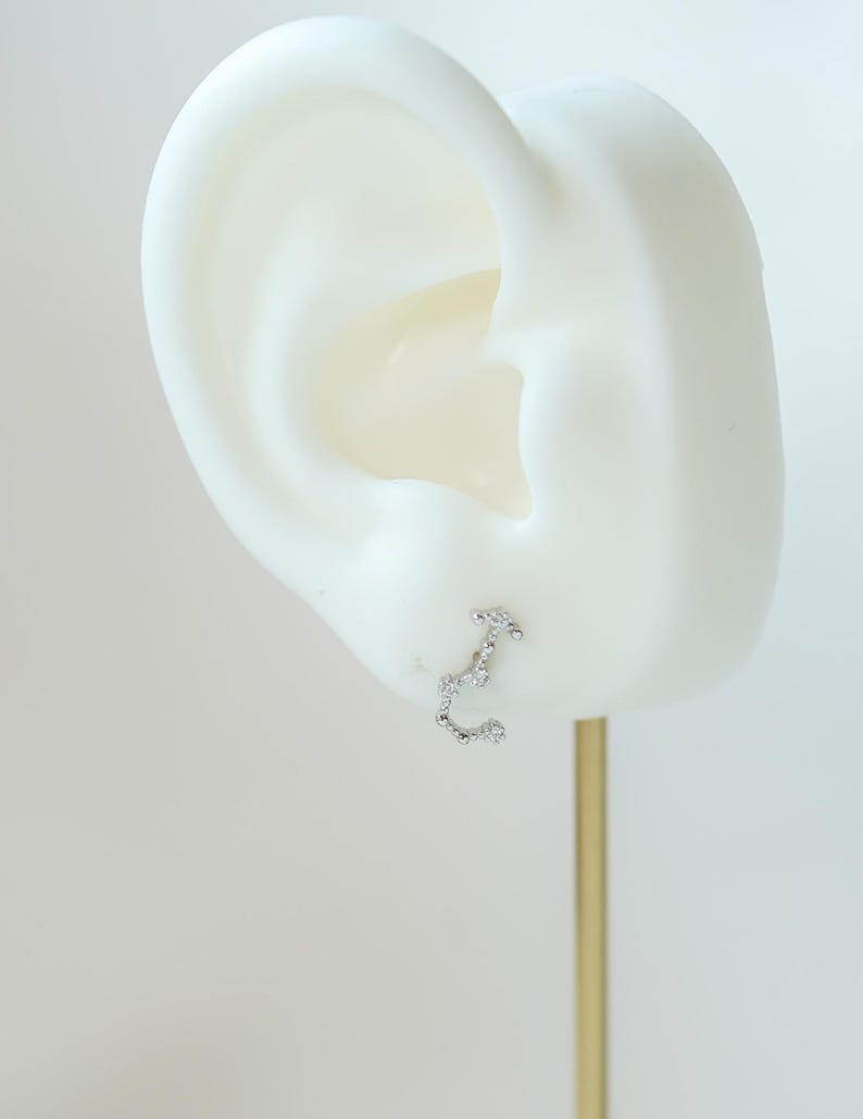 May include: A silver stud earring shaped like the Scorpio constellation, adorned with small, clear gemstones. The earring is presented on a white ear-shaped display against a pale backdrop. The earring is attached to a gold-coloured post.