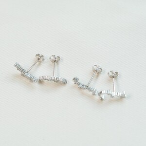 May include: Two pairs of silver stud earrings. Each earring has a linear design with multiple small, clear gemstones. The earrings are presented on a white surface, highlighting their delicate and sparkling appearance. These earrings are suitable for everyday wear.