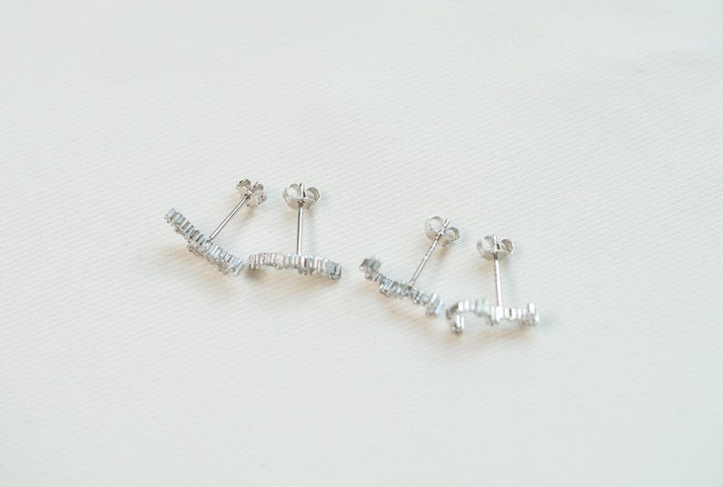May include: Two pairs of silver stud earrings. Each earring has a linear design with multiple small, clear gemstones. The earrings are presented on a white surface, highlighting their delicate and sparkling appearance. These earrings are suitable for everyday wear.