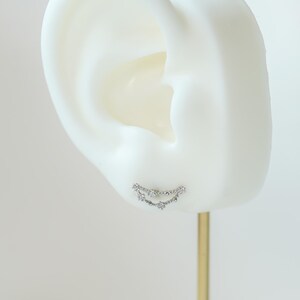 May include: A silver ear cuff earring with a curved design, set with small, sparkling gemstones. The earring is displayed on a white ear model against a neutral background.