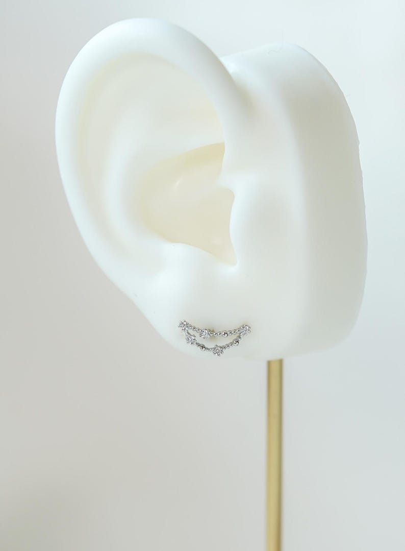 May include: A silver ear cuff earring with a curved design, set with small, sparkling gemstones. The earring is displayed on a white ear model against a neutral background.