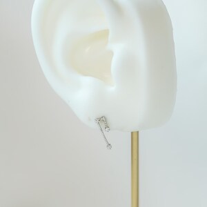 May include: A silver earring in the shape of a musical note, adorned with three clear, round gemstones. The earring is presented on a white ear-shaped form against a plain backdrop. The earring is attached to a gold-coloured metal stand.