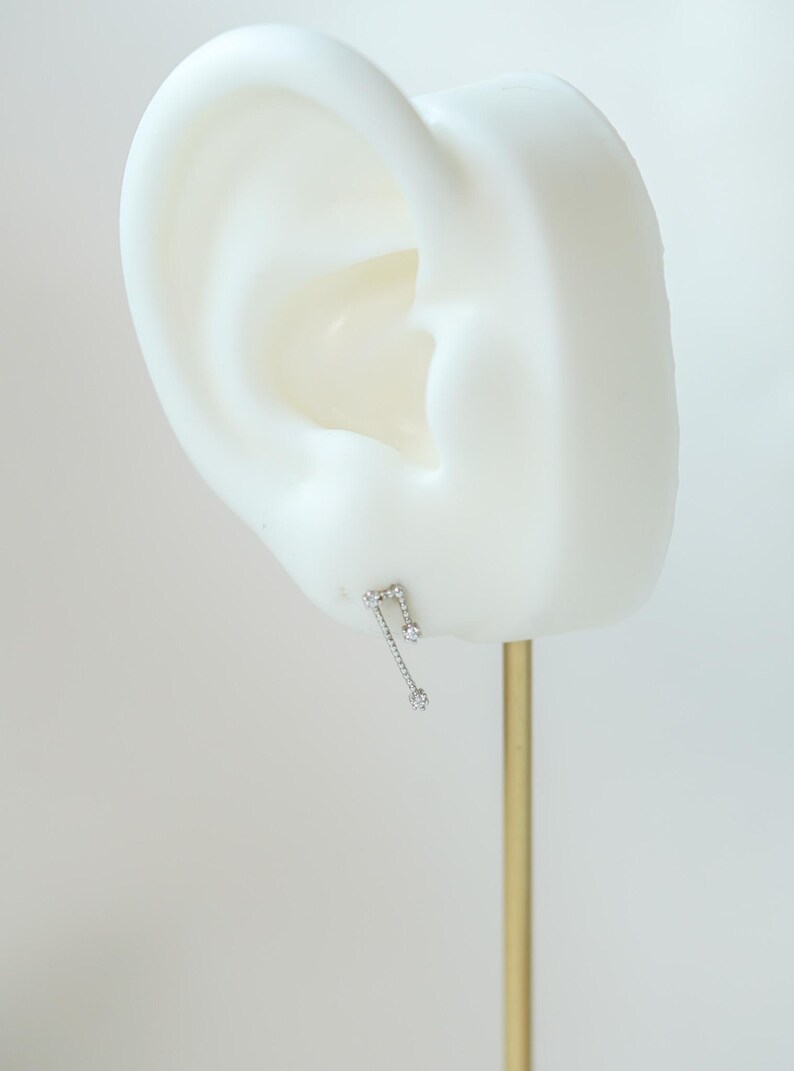 May include: A silver earring in the shape of a musical note, adorned with three clear, round gemstones. The earring is presented on a white ear-shaped form against a plain backdrop. The earring is attached to a gold-coloured metal stand.