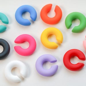 Könnte beinhalten: Eine Sammlung farbenfroher, C-förmiger Wein-Charm-Anhänger in verschiedenen Farbtönen, darunter Türkis, Blau, Orange und Grün. Weitere Farben sind Schwarz, Pink, Gelb und Rot. Diese Anhänger dienen zur Kennzeichnung von Weingläsern.