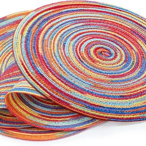 May include: Two colorful, round placemats with a spiral design. The placemats feature concentric rings of red, orange, yellow, blue, and green. The placemats are made of a woven material and are suitable for dining.