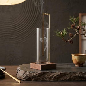 May include: A clear glass incense burner with a brass holder and a wooden base, featuring a thin incense stick emitting smoke. Beside it are three dark ceramic plates, a gold-toned metal tool, a small ceramic cup, and a miniature pine tree.