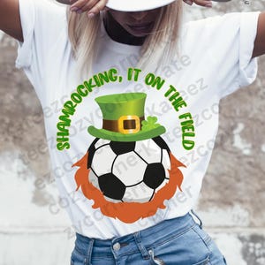 May include: White t-shirt with a green shamrock hat on a soccer ball with an orange beard. The text on the shirt reads "Shamrockin, It on the Field."