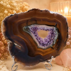 May include: A polished agate geode slice, showcasing concentric bands of brown, purple, and white. The center reveals a sparkling amethyst crystal formation. The geode is displayed on a silver metal stand, perfect for home decor or crystal enthusiasts.