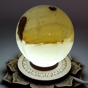 May include: A decorative lamp featuring a glowing, spherical light source with brown inclusions, resting on a wooden base. The base is intricately carved with a floral design. The lamp emits a soft, warm light.