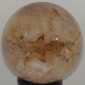 May include: A spherical, translucent crystal ball with a light brown and cream color scheme. The crystal ball has a rough, textured surface with a large, open cavity revealing the interior. The sphere is displayed on a black stand.