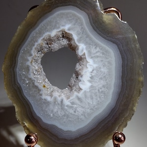 May include: A circular agate slice featuring concentric bands of gray, white, and translucent brown tones. The center contains a hollow geode cavity lined with small, sparkling white quartz crystals. The stone is supported by a copper-colored metal stand.