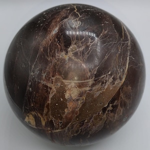 May include: A polished, spherical gemstone with a deep brown base color. The surface is marked with intricate patterns of lighter brown and cream-colored veins, creating a marbled effect. The sphere is smooth and reflective.