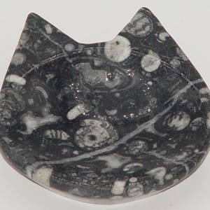 May include: A black and white stone dish shaped like a cat's head. The dish has a rounded bottom and two pointed ears at the top. The stone has a unique pattern of circular and linear markings.