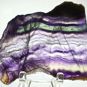 May include: A polished slice of fluorite mineral featuring horizontal bands of deep purple, translucent white, and pale green. The stone has an irregular, organic shape with a smooth, flat surface and natural jagged edges along the bottom.