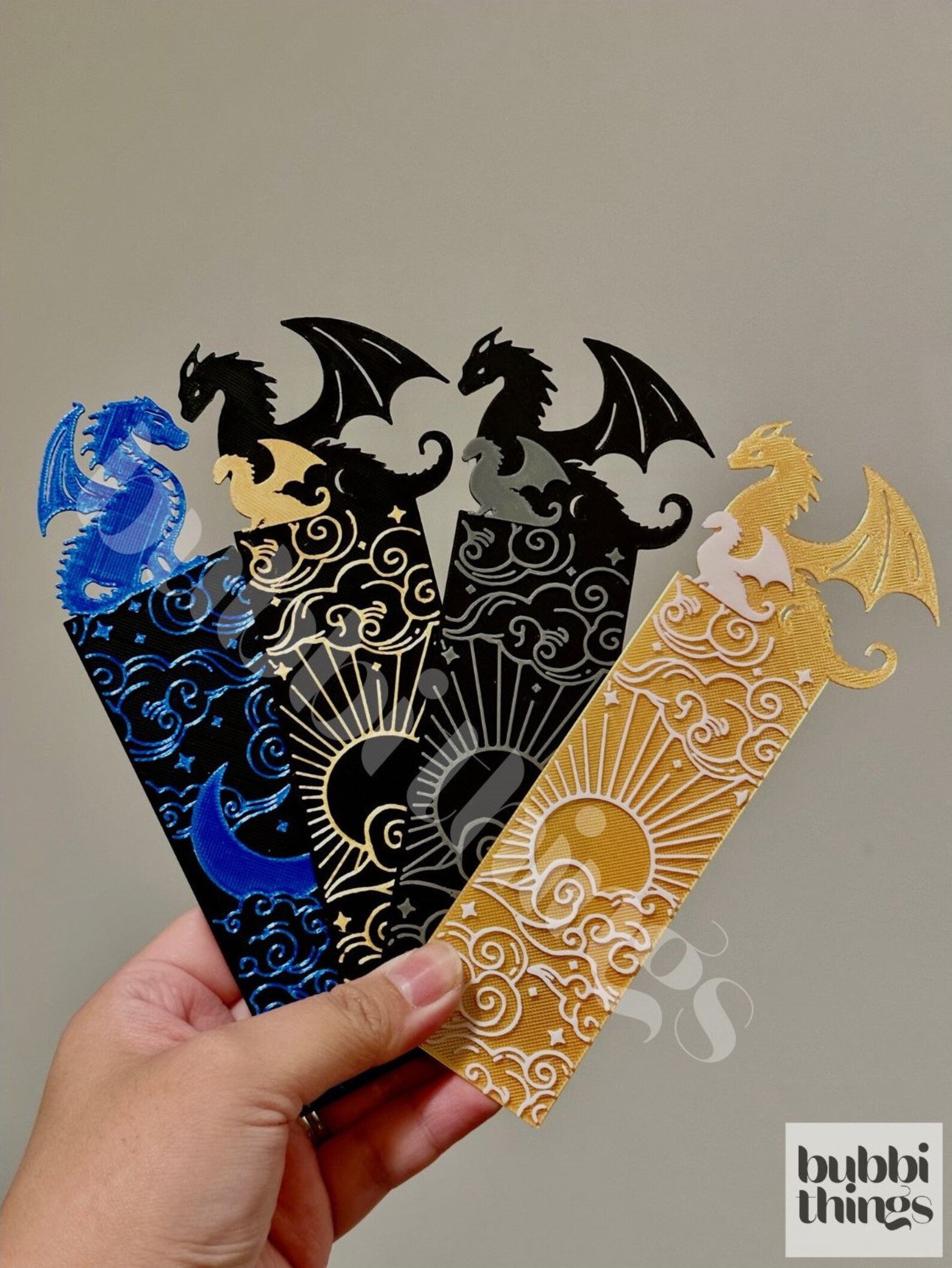 Four 3D-printed bookmarks of different colors (right to left: blue and black, gold and black, silver and black, gold and white). They have dragons at the top, and the middle to bottom are rectangular with thin sun/moon and cloud designs.