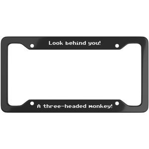 May include: Black license plate frame with white text: "Look behind you!" at the top and "A three-headed monkey!" at the bottom. The frame is rectangular with rounded corners. A humorous car accessory.