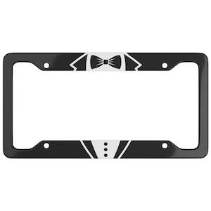 Peut inclure: Cadre de plaque d'immatriculation noir avec un motif de smoking. Le haut présente un nœud papillon et des revers blancs, tandis que le bas comporte des boutons blancs. Le cadre est rectangulaire.