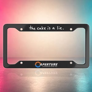 May include: Black license plate frame with the text "the cake is a lie." The frame also features the Aperture Laboratories logo. The frame is rectangular with rounded corners and mounting holes.