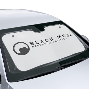 May include: A white car sunshade displaying the text "BLACK MESA RESEARCH FACILITY" in black. It also features a black circular logo with a partial square inside. The sunshade is fitted in a silver car.