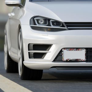 K&ouml;nnte beinhalten: Ein wei&szlig;es Auto mit einem dekorativen Kennzeichenrahmen. Der Rahmen hat einen schwarzen Hintergrund mit einem farbenfrohen Design mit roten, wei&szlig;en und grauen Elementen. Das Auto befindet sich auf einer Stra&szlig;e.