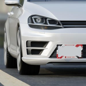 May include: A white car front bumper featuring a rectangular license plate frame with a red, black, and white graphic design. The frame displays the text "TRAUMA TEAM" in a stylized font, along with small geometric patterns and red accents.